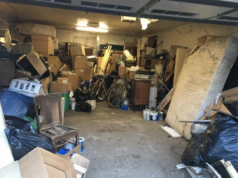 A cluttered garage packed with boxes, a mattress, bags and old furniture, the before photo